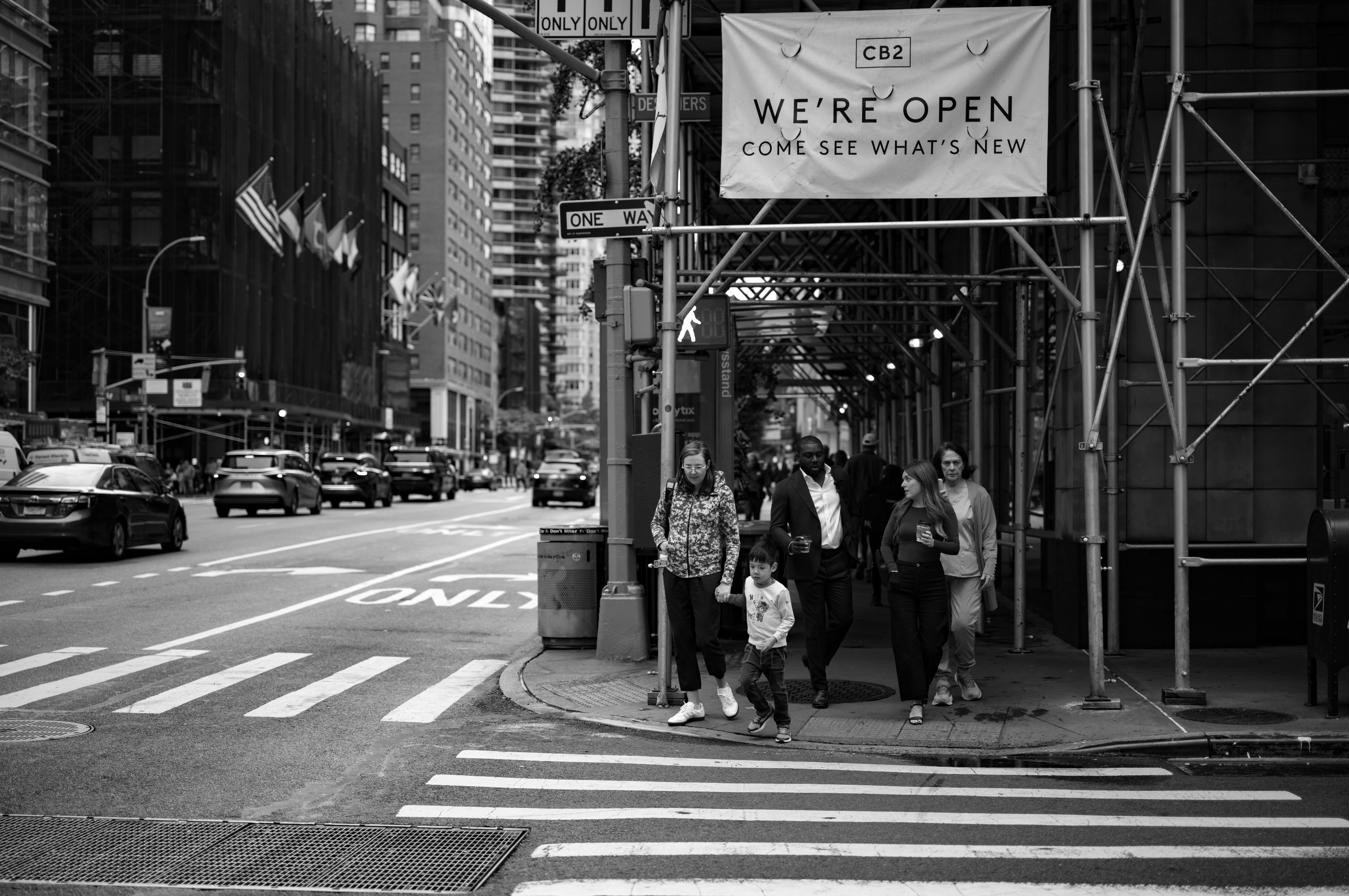 Pedestrians crossing a midtown Manhattan intersection past construction scaffolding, with a 'WE'RE OPEN' banner above